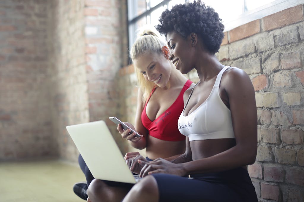 Two women in sportswear enjoy leisure time, sharing a laptop and smartphone in a gym setting against a brick wall.